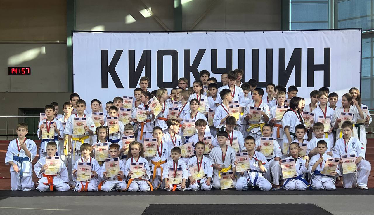 Дети и подростки в каратэ-ги с цветными поясами позируют с дипломами и медалями на фоне баннера «Киокушин» после турнира в спортивном зале.
